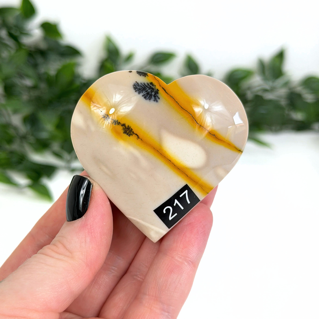 Heart-shaped mookaite with yellow and black patterns held in a hand against a blurred green background
