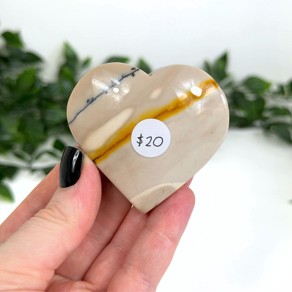 Heart-shaped mookaite crystal with a price tag held by a hand against a blurred green plant background