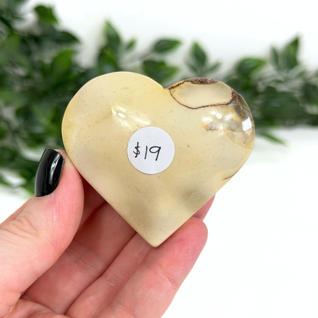 Heart-shaped mookaite stone held in a hand with a price tag sticker, against a blurred green leafy background.