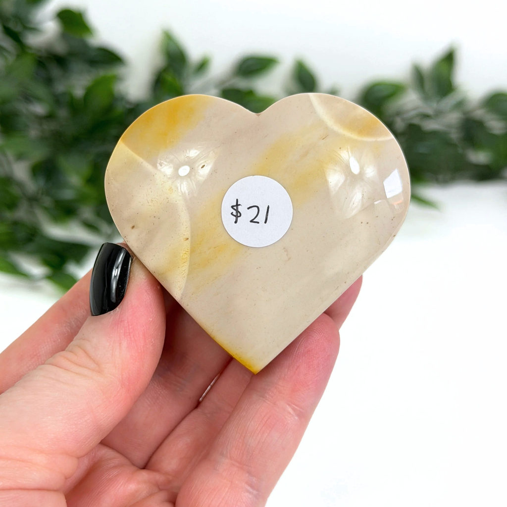Heart-shaped mookaite crystal held in a hand with a price tag sticker, greenery in the background